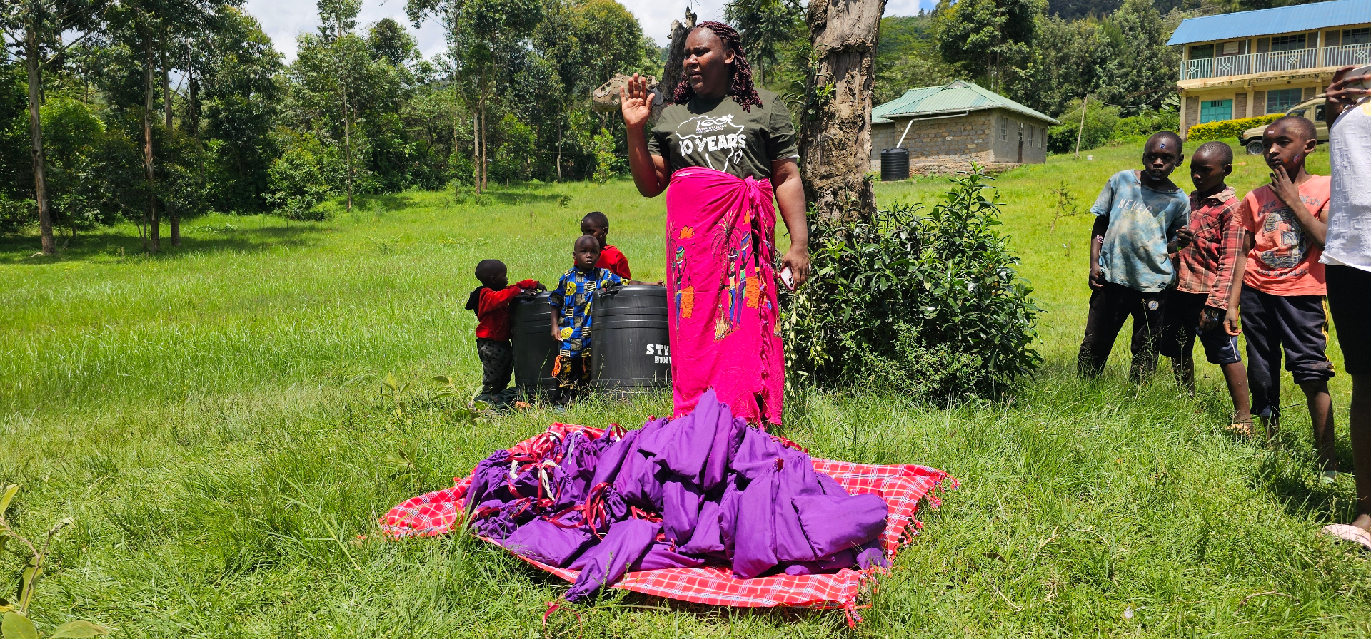 100 Humanitarians community leader standing beside a pile of purple HopeKit bags in a Kenyan hillside field, ready for distribution to women and girls