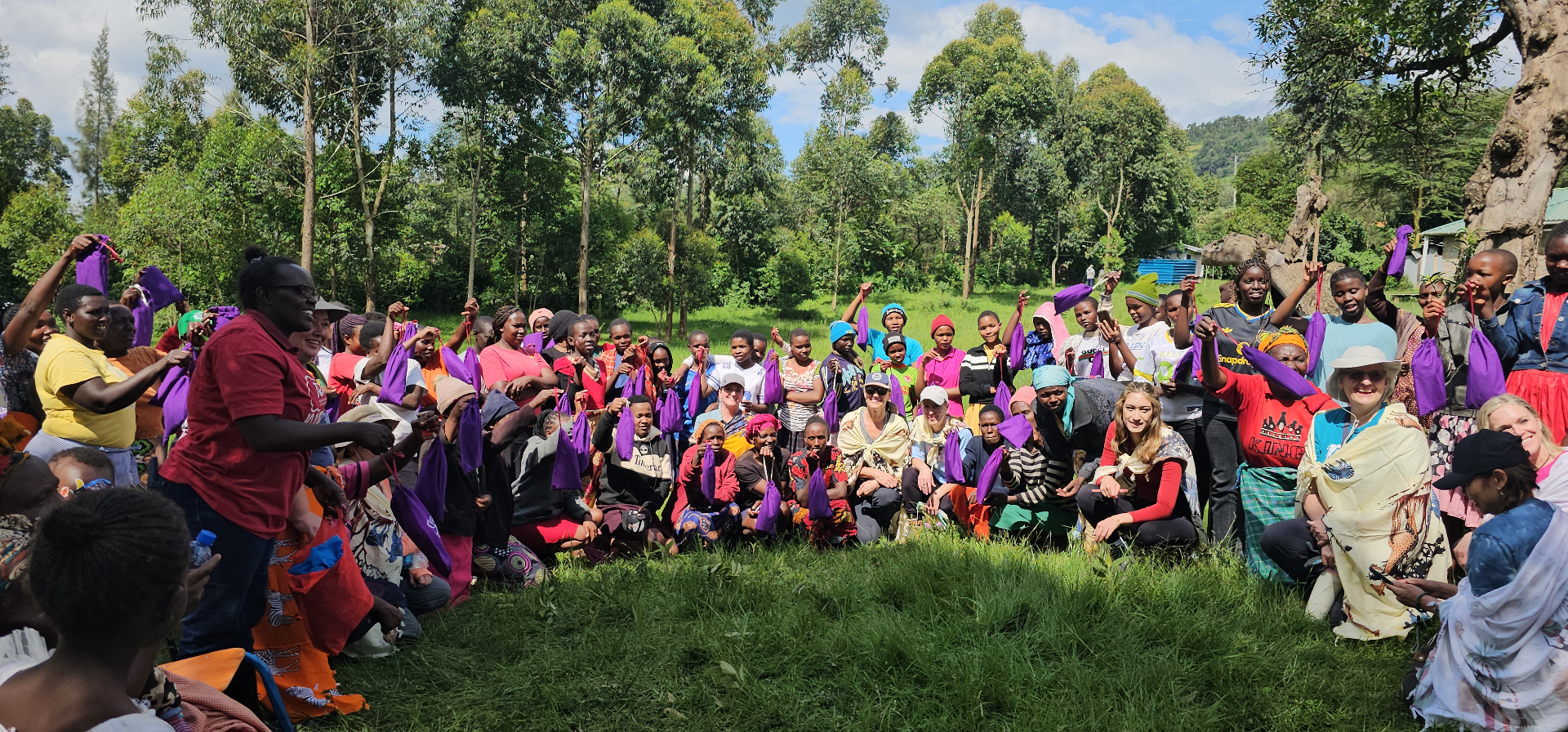 Large group of women and girls in a forest clearing raising purple HopeKit bags in celebration at a 100 Humanitarians distribution event in Kenya