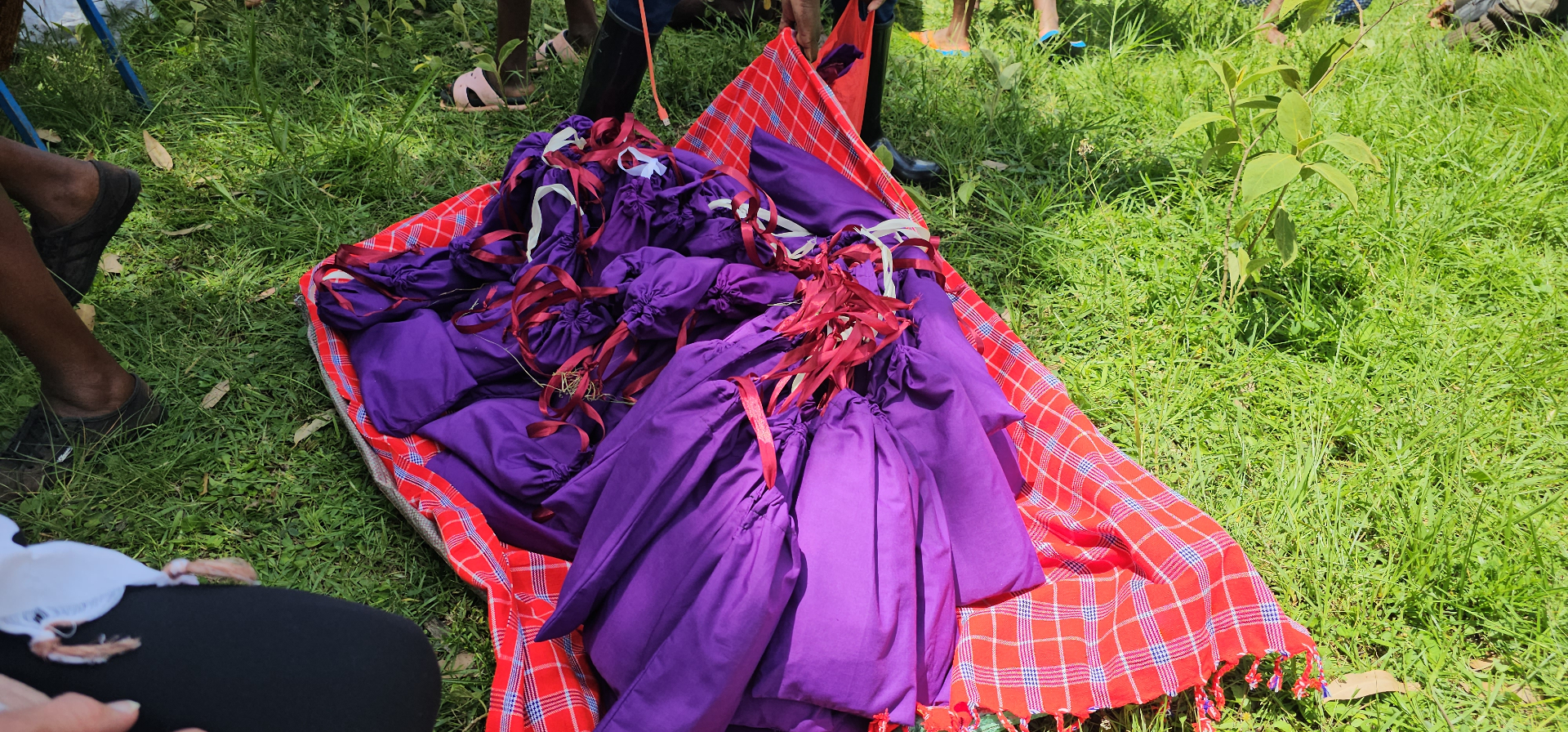 A pile of purple HopeKit drawstring bags with red ribbons resting on a traditional red Maasai shuka in the grass
